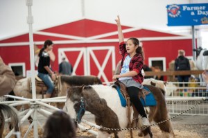 A young girl on a pony rides near a red barn